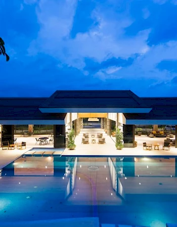 A luxurious indoor-outdoor space with a large, illuminated pool in front of a modern building with lounge chairs and tables.