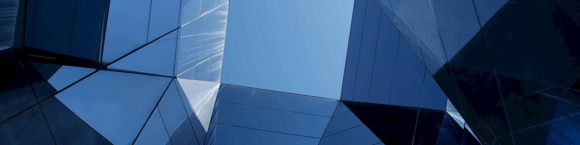 View of geometric glass structures forming an abstract pattern with a central opening to the sky, producing a reflective and mirrored effect.