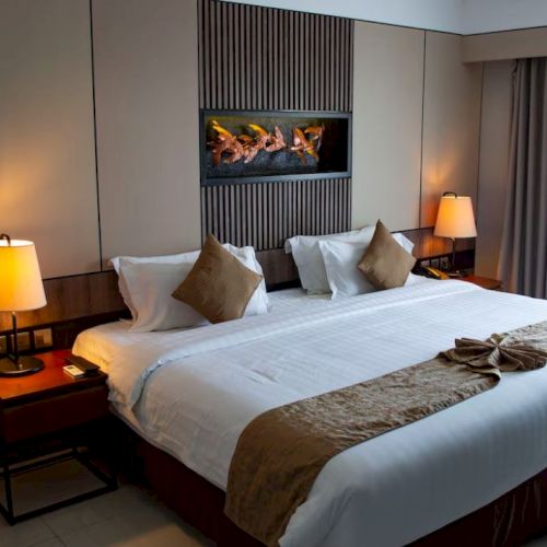 The image shows a neatly arranged hotel room with a large bed, two bedside tables with lamps, beige and brown decor, and a modern aesthetic.
