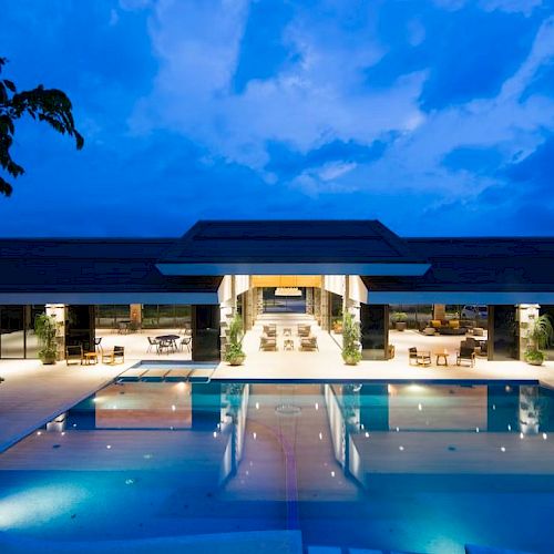 The image shows a luxurious open-air building at dusk, featuring a large swimming pool surrounded by lounge chairs and illuminated by ambient lighting.