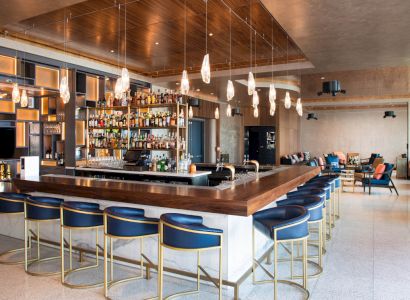 A modern bar with sleek blue stools, hanging pendant lights, a well-stocked liquor shelf, and a cozy seating area in the background.