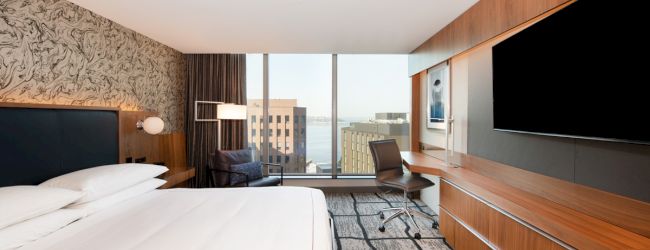 A modern hotel room with a king-sized bed, large flat-screen TV, desk, chair, and window with a city view overlooking buildings and water.