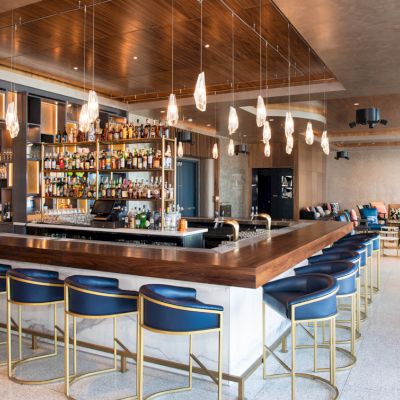 Modern bar with a wooden counter, blue bar stools, hanging lights, and a well-stocked drink shelf, plus a cozy seating area in the background.