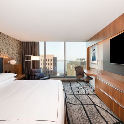 A modern hotel room features a king-size bed, wall art, floor-to-ceiling windows, a cityscape view, a large TV, workspace, and contemporary furnishings.
