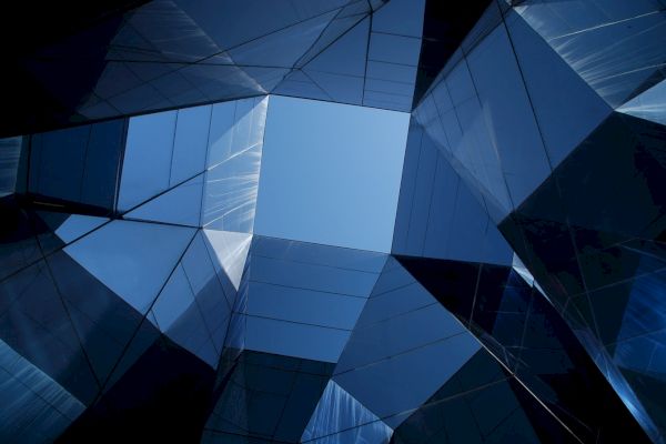 Looking up through a geometric arrangement of reflective, angular glass structures, revealing a portion of clear blue sky at the center.