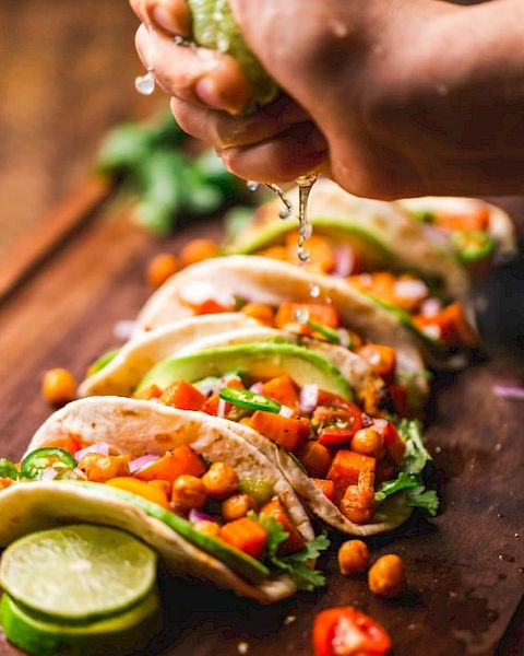 A hand is squeezing lime juice over a row of freshly prepared tacos filled with vegetables, chickpeas, avocado slices, and garnished with cilantro.