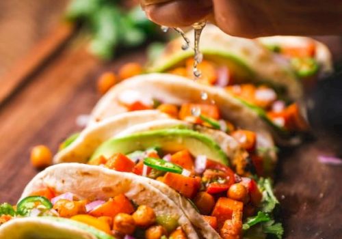 A hand is squeezing lime juice over a row of freshly prepared tacos filled with vegetables, chickpeas, avocado slices, and garnished with cilantro.
