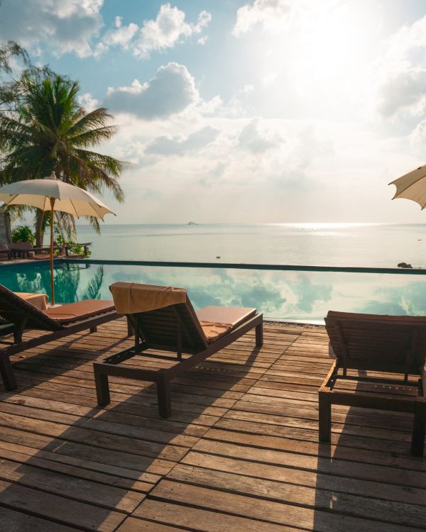 Lounge chairs and umbrellas on a wooden deck overlook a serene infinity pool with a tropical ocean view under a bright sky.