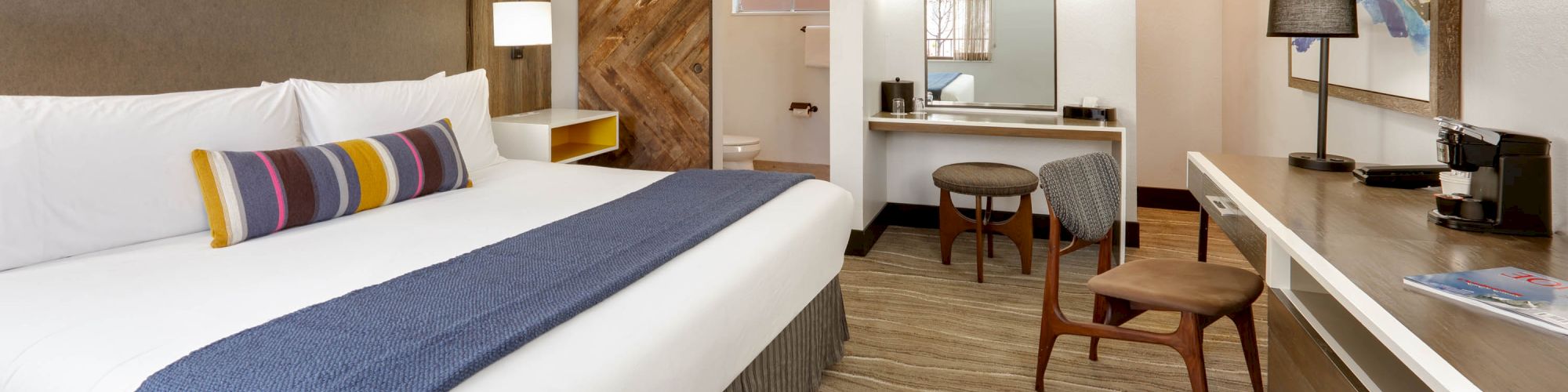 This image shows a modern hotel room with a king-sized bed, a desk, a chair, and a bathroom with a sliding barn door.