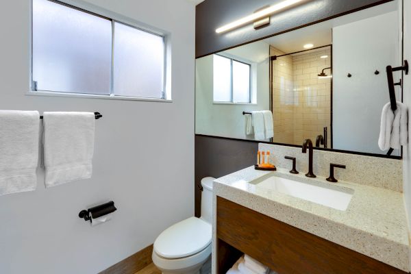 A modern bathroom with a large mirror, white countertop, black faucet, toilet, towels, and a glass-enclosed shower with subway tiles.