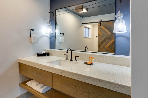 A modern bathroom with a large mirror, single sink, dark fixtures, hanging lights, and wooden cabinetry. Shelves below hold white towels.