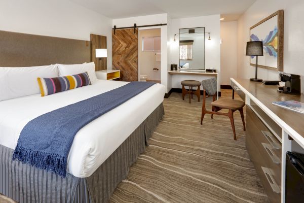A modern hotel room with a king-sized bed, blue throw, wood accents, desk, mirror, coffee maker, and a sliding barn door leading to the bathroom.