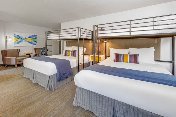 The image shows a hotel room with two double beds, each with a bunk overhead, a small seating area, and modern decor including colorful pillows.