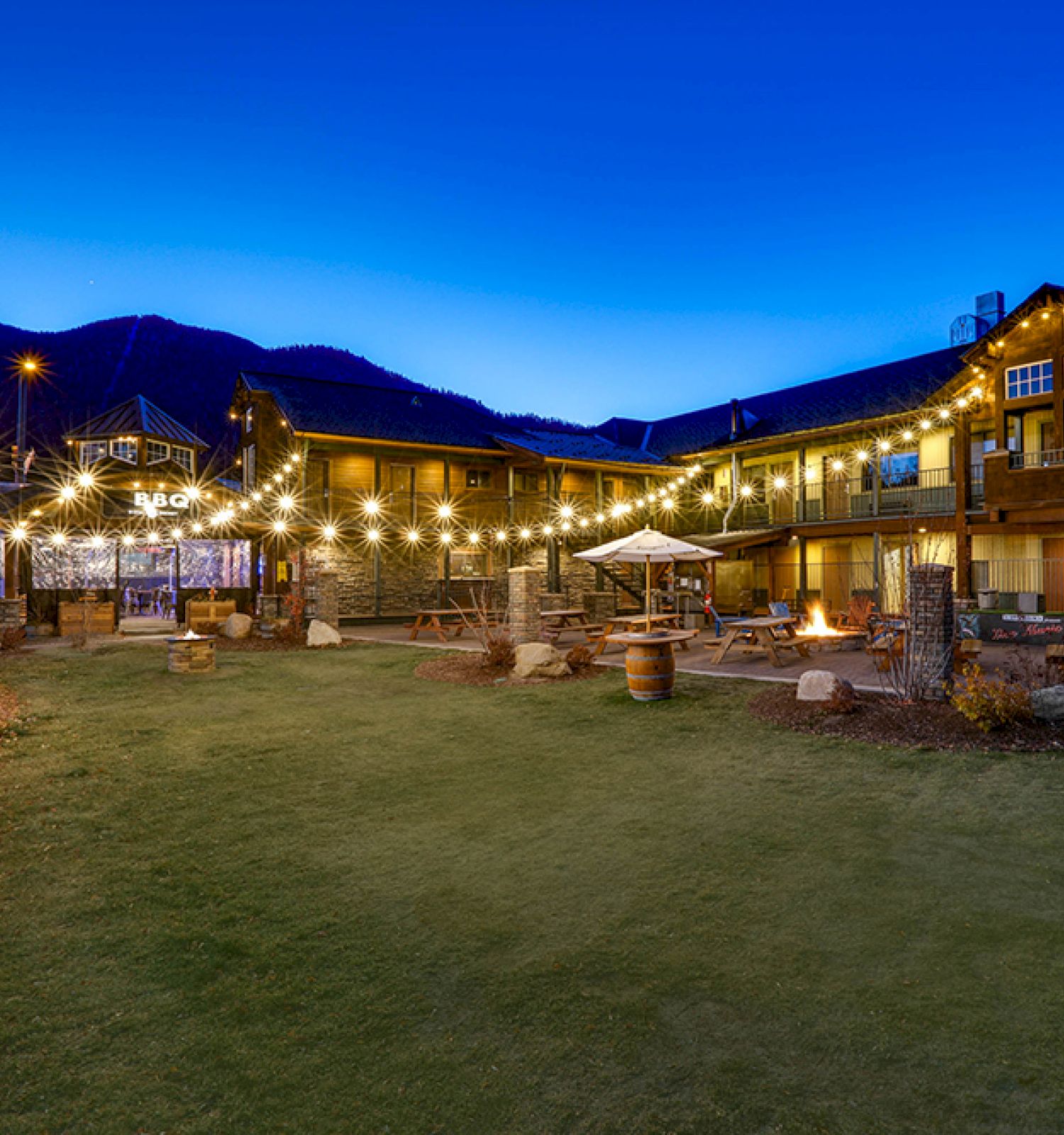 A cozy outdoor area of a lit-up hotel or lodge during the evening, featuring string lights, a green lawn, and mountains in the background.