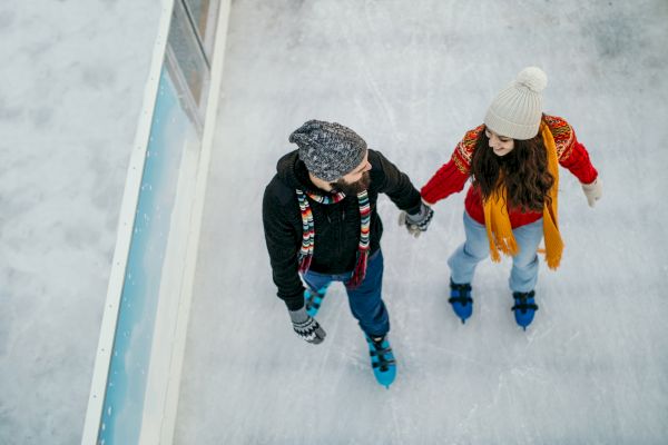 Two people are ice skating holding hands, both wearing winter clothing including hats and scarves, with a barrier on the left.