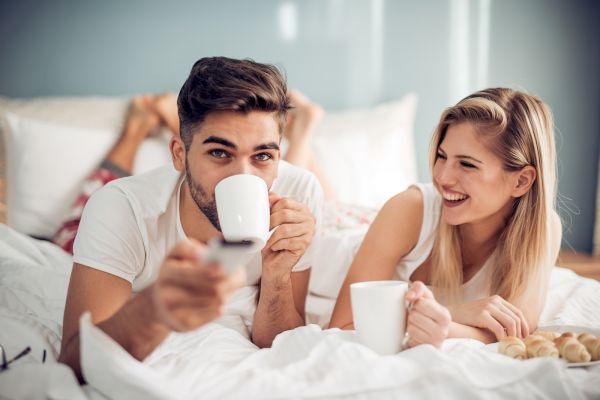 A man and woman are lying in bed, both holding white mugs, smiling and enjoying a cozy moment together, with pastries on the side.