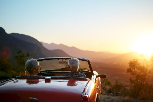 Two people are sitting in a red convertible, enjoying a scenic mountain sunset.