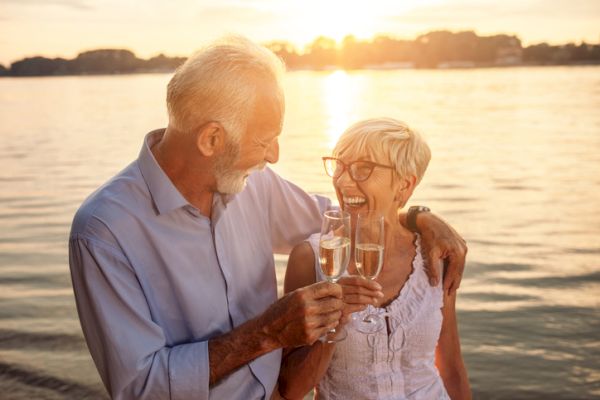 An older couple is enjoying a sunset by the water, smiling and toasting with champagne glasses, exuding happiness and love.
