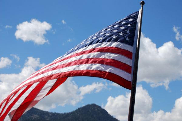 An American flag is waving in the wind with a clear blue sky and a mountainous landscape in the background.