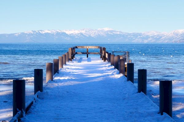 A snow-covered wooden pier stretches out into a calm, blue lake with mountains in the background under a clear sky, creating a serene scene.