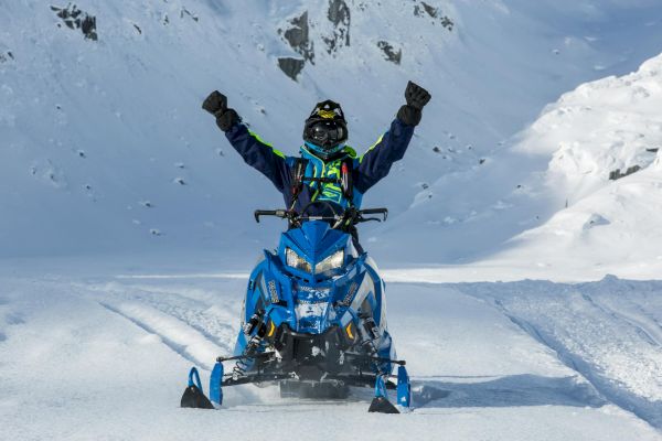 A person riding a blue snowmobile is raising their arms triumphantly on a snowy landscape with mountains in the background.