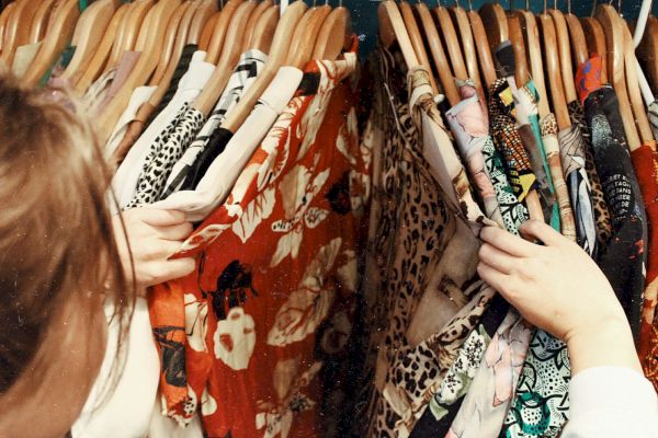A person is browsing through a rack of various colorful clothes on wooden hangers, with patterns such as florals and animal prints.