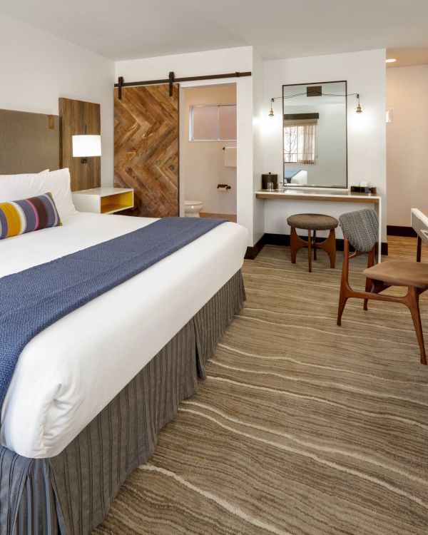 A well-decorated hotel room with a king-sized bed, a wooden sliding door, a desk, a chair, and a mirror, featuring modern amenities and decor.
