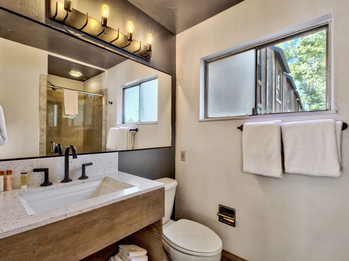 A modern bathroom with a large mirror, sink, toilet, two towel racks with white towels, and a window with a view of trees and adjacent building.