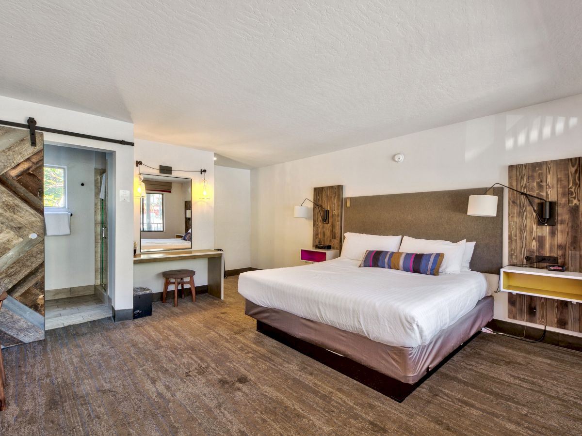 A modern hotel room with a king-sized bed, wood accents, sliding barn door, desk, and a bathroom with a glass shower enclosure is shown.