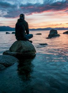 A person is sitting on a rock in the middle of a calm body of water, watching a colorful sunset or sunrise, surrounded by other rocks.