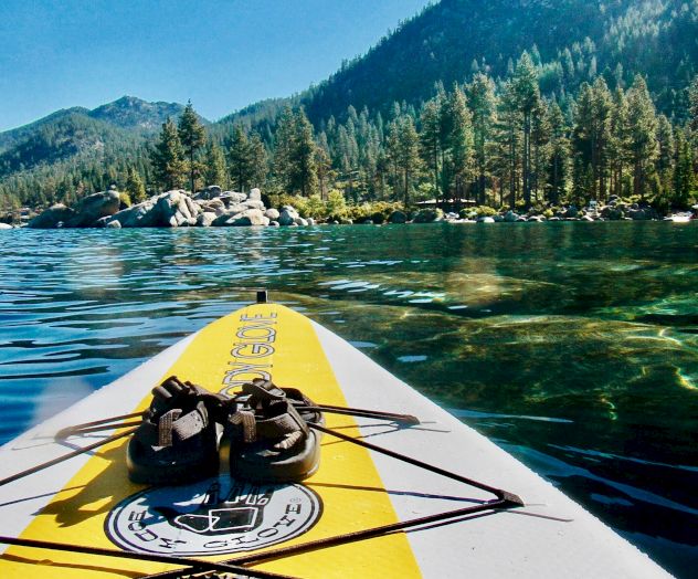 A paddleboard with shoes on it is floating on a calm lake, surrounded by trees and mountains under a clear blue sky.