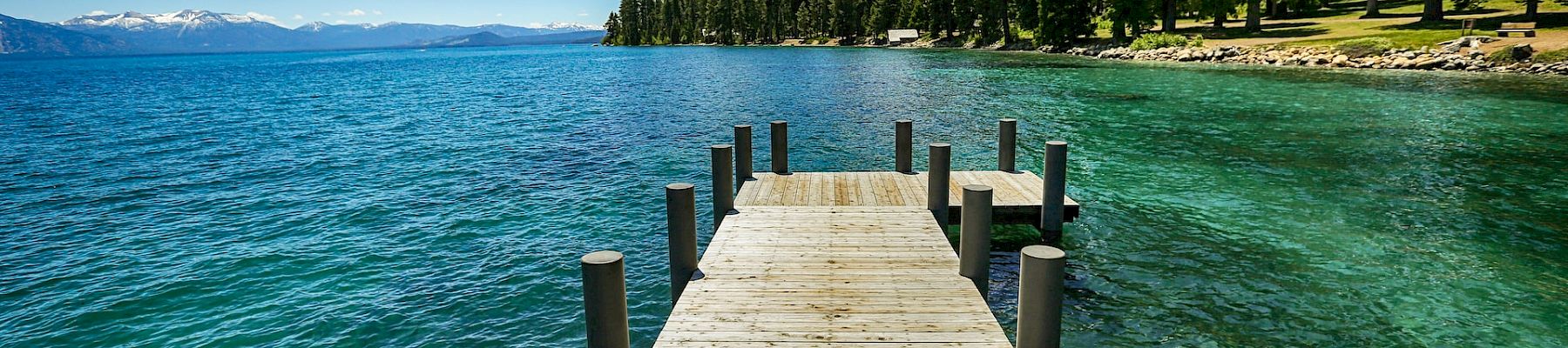 A wooden pier extends into a clear blue lake surrounded by tall pine trees on a sunny day, with mountains visible in the distance.