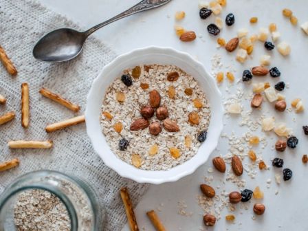 A bowl of muesli with assorted nuts, dried fruits, and oats, surrounded by scattered pretzel sticks and a spoon on a textured cloth.