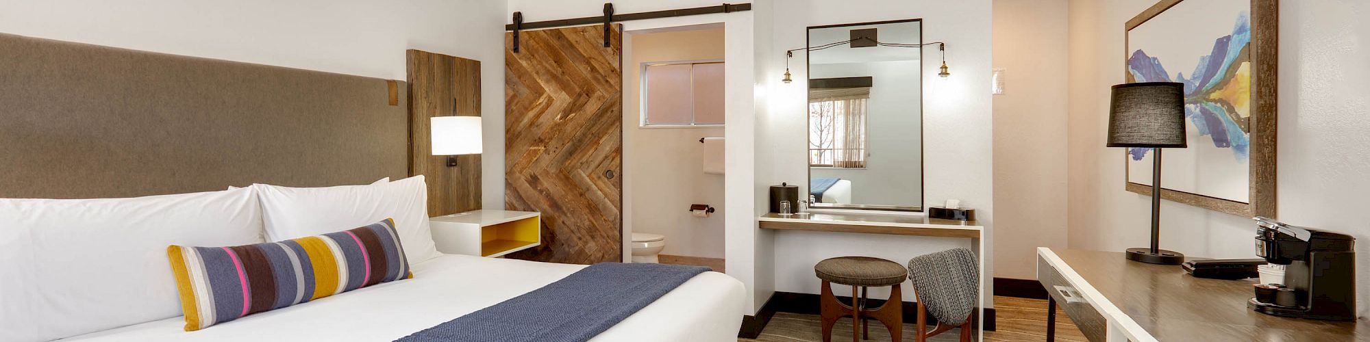 A modern hotel room with a large bed, blue throw, work desk, chair, vanity area, and bathroom with sliding barn door and contemporary decor.