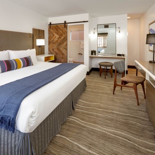 The image shows a modern hotel room with a king-sized bed, nightstands, a desk, a mirror, a chair, and a bathroom with a wooden sliding door.