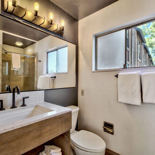 A modern bathroom with a large mirror, black fixtures, a shower area, a toilet, toiletries, and towels on a rack by the window.
