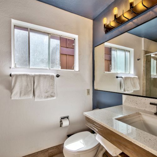 A modern bathroom with a large mirror, window, white countertop, black fixtures, and towels on a rack.