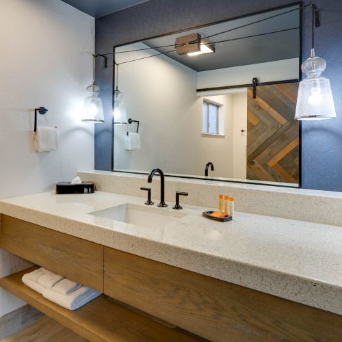 This image shows a modern bathroom with a large mirror, a single sink with a black faucet, hanging lights, and wooden cabinetry below the countertop.