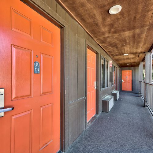An outdoor hallway with several orange doors labeled with room numbers, gray walls, and railings. Light fixtures are mounted on the wooden ceiling.