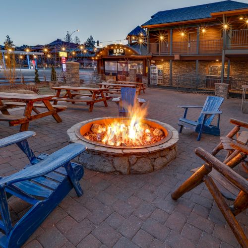 An outdoor seating area with wooden and Adirondack chairs surrounding a fire pit, nearby is a BBQ restaurant lit with string lights in the evening.