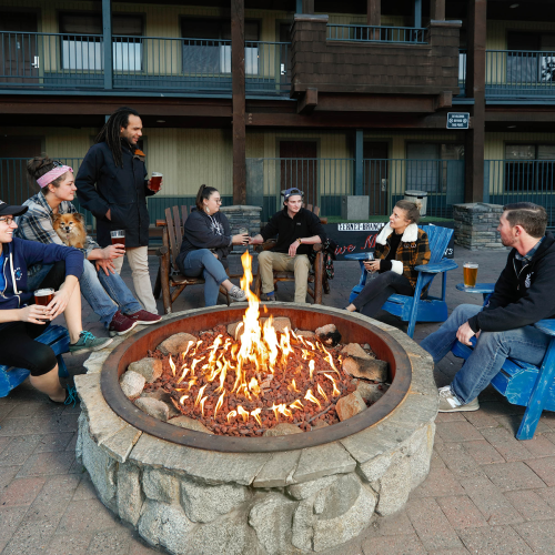 A group of people sits around a fire pit in outdoor chairs, casually talking and holding beverages.