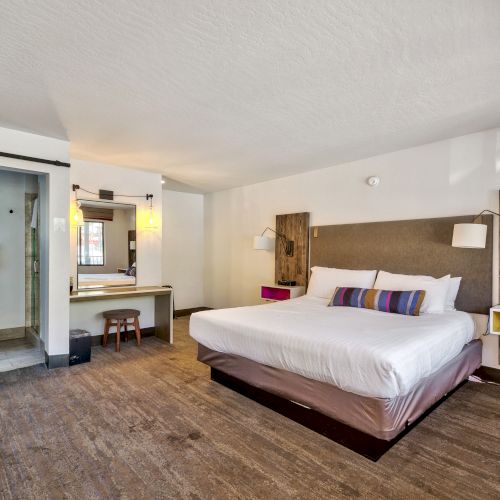 A modern hotel room featuring a large bed with colorful pillows, a workspace, wooden decor, and an open bathroom with a sliding barn door.