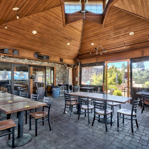 An outdoor dining area with wooden ceilings, stone walls, and large windows. The space is furnished with wooden tables and chairs in a casual setting.