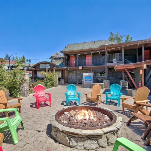 A cozy outdoor seating area with colorful Adirondack chairs arranged around a stone fire pit, surrounded by rustic buildings under a clear blue sky.