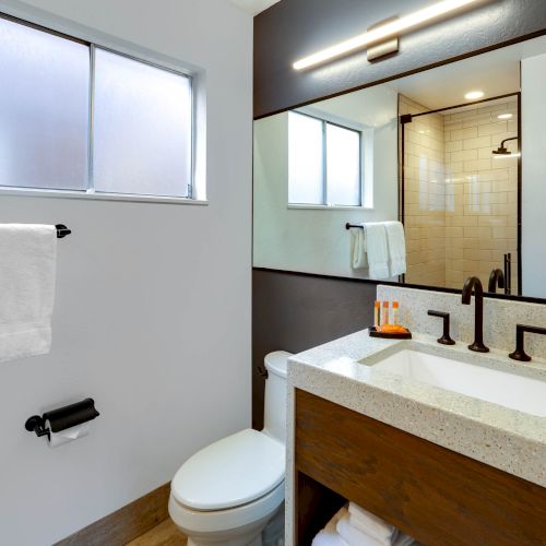 A modern bathroom with a large mirror, sink, towels, toilet, and a glass shower enclosure, featuring sleek fixtures and a minimalist design.