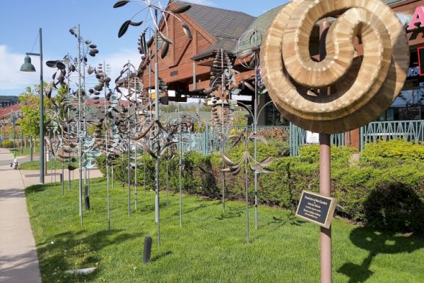 The image shows a sculpture garden with metal wind sculptures on a grassy area, a wooden circular sculpture, and buildings in the background.
