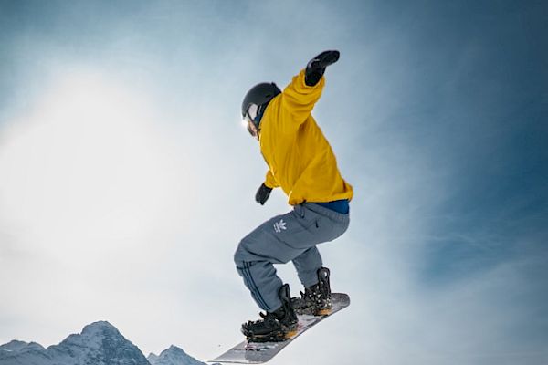 A snowboarder in a yellow jacket is performing a trick on a snowy rail with mountains in the background, under a clear sky.
