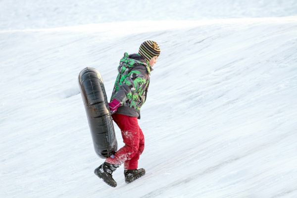 A person in winter clothing carries a black sled or inner tube up a snowy slope, preparing for a downhill ride.