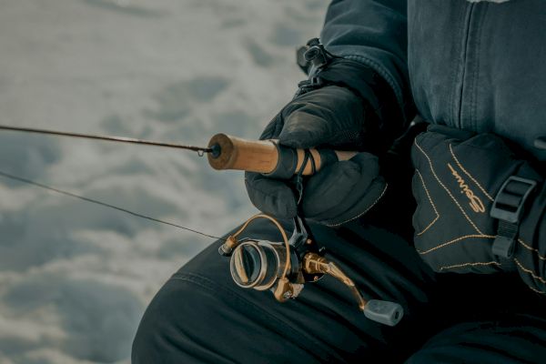 A person in winter gear holds a fishing rod, appearing to be ice fishing in a snowy environment.