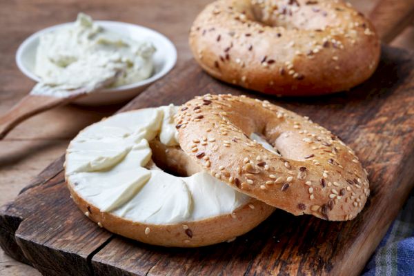 Two sesame bagels on a wooden board, one with cream cheese spread, next to a bowl of extra cream cheese and a wooden knife.